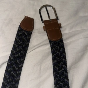 Mörkblått flätat bälte med skinn - Snyggt flätat bälte i mörkblått och grått med bruna skinn-detaljer vid spännet och änden. Bältet har ett klassiskt silverfärgat spänne och passar perfekt till jeans eller chinos. Den flätade designen ger en cool och avslappnad vibe. Inga skador på bältet. 