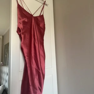 Rosa långklänning Linn Ahlborg x Rebecca Stella - Snygg långklänning i glansig rosa satin från Linn Ahlborg x Rebecca Stella. Klänningen har tunna axelband som korsas i ryggen och en figurnära passform. Perfekt för dig som vill sticka ut med en elegant och trendig look.