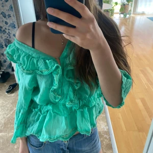 Mintgrön/ turkos volangblus med v-ringning - Supersöt turkos blus med volanger och offshoulder-look. Blusen har v-ringning och långa ärmar. Perfekt för dig som gillar romantiska detaljer och en luftig känsla.