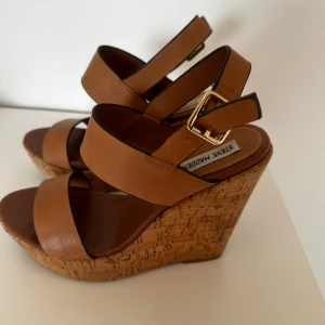 Kilklack Steve Madden - Snygga sandaler med breda bruna remmar i skinn och justerbart spänne i guld. Korkklädd platåklack ger extra höjd och sommarkänsla. Perfekta för dig som vill ha både stil och komfort. Rund öppen tå och stabil plattform.   Nypris: 1200kr