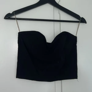 Svart bandeau tubtopp - Säljer en svart bandeau tubtopp med hjärtformad urringning och transparenta axelband. Toppen är croppad och har en stilren, minimalistisk look som passar perfekt till höga jeans eller kjol. Enkel att matcha och supertrendig för dig som gillar en clean vibe.
