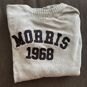 Ljusgrå tröja Morris L - Snygg ljusgråtröja från Morris i storlek L. Tröjan har broderad text 'Morris 1968' framtill och Union Jack-patch på ärmen. Ribbstickade muddar och krage ger en klassisk look. Perfekt för dig som gillar preppy stil.
