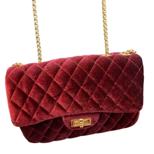 Mörkröd quiltad axelväska i sammet - Supersnygg mörkröd axelväska i mjuk sammet med quiltat mönster och guldfärgad kedja. Väskan har ett klassiskt lock med vridspänne i guld. Perfekt för att lyfta vilken outfit som helst.