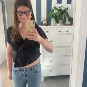 Svart omlotblus med kort ärm - Snygg svart omlotblus med korta ärmar och croppad passform. Blusen har en djup v-ringning och knytdetalj i sidan som ger en cool och trendig look. Perfekt att matcha med jeans eller kjol för en stilren outfit.