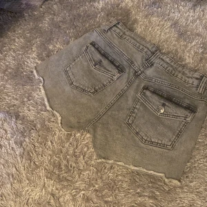 Grå jeansshorts med rå kant - Snygga grå jeansshorts med rå, fransig kant och klassiska fem fickor. Shortsen har bälteshällor och knäppning fram, samt bakfickor med lock och knapp. Perfekta för en chill sommarlook.