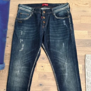Mörkblå jeans med lönnlöv från Dsquared2 - Snygga mörkblå jeans från Dsquared2 med slitningar och tvättade detaljer framtill. Bakfickan har en röd broderad lönnlöv. Klassisk femficksmodell med smal passform och knappgylf. Perfekta för dig som gillar streetstyle med en twist. Strlk L32