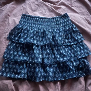 Blå volangkjol med mönster - Supersöt blå kortkjol med volanger och elastisk midja. Kjolen har ett stilrent mönster i vitt och blått och är perfekt för dig som gillar en lekfull och trendig look. Lätt och luftig, passar perfekt till varmare dagar.