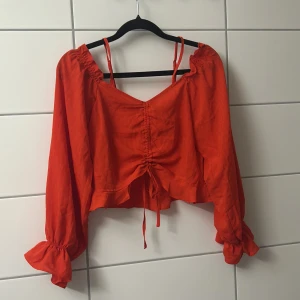 Röd croppad blus med snörning - Trendig röd blus med croppad passform, långa puffiga ärmar och snygg snörning framtill. Blusen har volangdetaljer vid ärmsluten och smala axelband. Perfekt för dig som vill sticka ut med färg och coola detaljer.