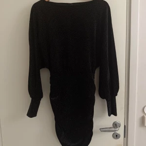 Svart glittrig kortklänning - Snygg svart kortklänning med glittriga detaljer och långa ärmar. Klänningen har markerad midja och lätt puffiga ärmar med breda muddar. Perfekt för dig som vill sticka ut med en elegant och trendig look.