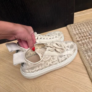 Chloé spets-sneakers i beige - Unika sneakers från Chloé i beige spets med coola broderade detaljer och tydlig logga på hälen. Skorna har snörning, platt vit sula och stora tygflikar bak. Perfekta för dig som vill sticka ut med en lyxig och feminin touch.