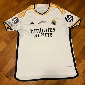 Real Madrid Bellingham Finaltröja 2024 - Real Madrid fotbollströja från Champions League-finalen 2024 i London, med Bellingham och nummer 5 på ryggen. Vit med svarta och gula detaljer, broderat klubbmärke, Adidas-logga och sponsortryck. Tillverkad i lätt polyester med korta ärmar och klassiska ränder på axlarna.