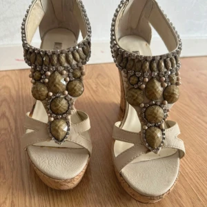 Crèmefärgd sandaler med kilklack och stenar - Unika crèmefärgad sandaler med hög kork-kilklack och plattformsula. Dekorerade med stora bruna och beige stenar, pärlor och glittrande detaljer framtill. Remmar i syntetmaterial och öppen tå ger en cool och somrig vibe. Perfekta för dig som vill sticka ut. Tyvärr har skotens små defekter