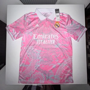 Adidas x Real Madrid fotbollströja  - Snygg rosa Real Madrid fotbollströja från Adidas med grått drakmönster och vit krage. Tröjan har korta ärmar, broderad klubbmärke och Adidas-logga samt tryckt sponsor. Tillverkad i lätt och ventilerande material, perfekt för match eller träning.