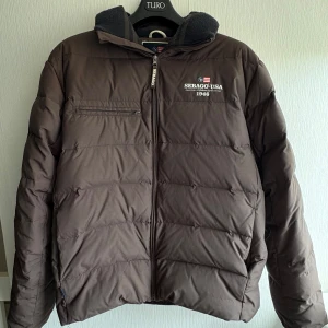 Brun pufferjacka från Sebago - Snygg brun pufferjacka från Sebago med broderad logga och amerikansk flagga på bröstet. Jackan har dragkedja framtill, en bröstficka med dragkedja och hög krage med stickad insida. Perfekt för kalla dagar och riktigt skön att bära.