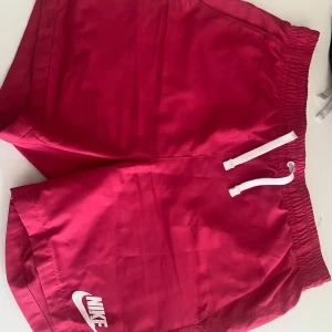 Rosa Nike shorts  - Snygga rosa shorts från Nike med vit logga på benet. De har elastisk midja med vit snörning och en bakficka med metallöljet. Perfekta för sommarens aktiviteter eller träning. Lätta och snabbtorkande material med meshfoder för extra komfort. Storlek S