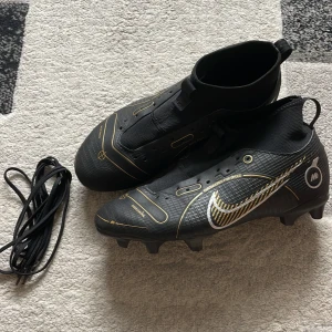 Nike fotbollsskor svart/guld strl 38.5 - Snygga svarta Nike fotbollsskor med guldiga detaljer och vit swoosh. Skorna har en strumpliknande passform, platt sula med dobbar och snöre medföljer. Perfekta för dig som vill sticka ut på planen med stilrena och sportiga skor.