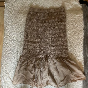 Beige volangkjol från H&M - Supersöt beige långkjol från H&M med smockad midja och volang nertill. Kjolen har en lätt glansig yta och är tillverkad i ett mjukt, flowy material som ger en snygg siluett. Perfekt att styla med en enkel topp eller stickad tröja.