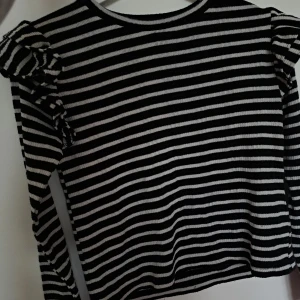 Randig svartvit tröja med volang Zara - Säljer en svart och vit randig tröja från Zara i storlek S. Tröjan har långa ärmar och coola volangdetaljer på axlarna. Materialet ger en skön känsla. Perfekt för dig som gillar en trendig och lekfull stil.