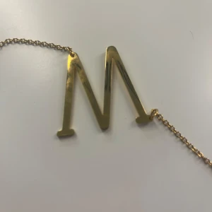 Guldigt halsband med bokstaven M - Stilrent halsband i guldton med en stor, blank bokstav M som hänge. Kedjan är tunn och har ett klassiskt lås. Perfekt accessoar för dig som vill ha något personligt och trendigt runt halsen. Aldrig använt. Nypris 320kr, ni kan få det gratis
