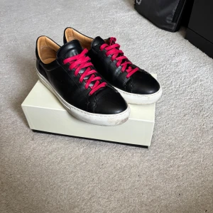 Svarta sneakers  - Stilrena svarta sneakers i skinn med vita sulor och röda snören som ger en snygg kontrast. Insidan är ljusbrun och skorna har en klassisk, rund tå. Perfekta för dig som gillar minimalistisk design med en färgklick. OBS! De är stora i storleken!