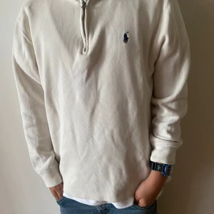 Vit half zip tröja från Polo Ralph Lauren - Vit tröja från Polo Ralph Lauren med half zip och blå broderad logga på bröstet. Tröjan är ribbstickad i bomull och har en klassisk, avslappnad passform. Perfekt för lager-på-lager och enkel att matcha med jeans eller chinos. Nypris 2699 säljes för 890.  large storlek 14 