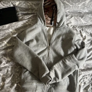 Grå Burberry hoodie med dragkedja - Säljer en grå hoodie från Burberry med klassiskt rutigt foder i huvan och broderad logga på bröstet. Tröjan har dragkedja framtill, två fickor och ribbade muddar. Perfekt för dig som gillar stilren streetwear med lyxig touch.