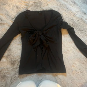 Svart långärmad topp med knyt Gina Tricot - Supersnygg svart långärmad topp från Gina Tricot i storlek XS. Toppen har en djup v-ringning och en stor knyt-detalj framtill som ger en trendig look. Perfekt att styla med jeans eller kjol för en cool vibe.