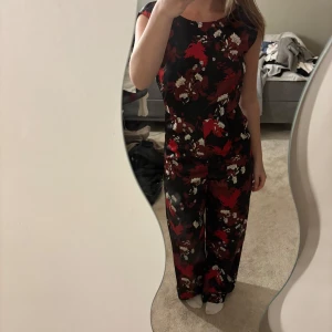 Blommig svart jumpsuit från ONLY - Supersnygg svart jumpsuit från ONLY med röda och vita blommor. Vida ben, öppen rygg och kan bäras off-shoulder. Sitter tajt upptill och har ett flowy fall nertill. Perfekt för dig som vill sticka ut med ett coolt mönster och skön passform.