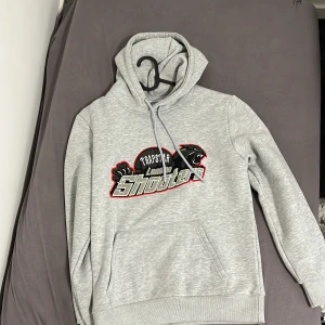 Grå hoodie Trapstar London Shooters - Snygg ljusgrå hoodie från Trapstar med broderad logga och texten 'London Shooters' framtill. Tröjan har huva med snörning, magficka och ribbade muddar. Perfekt för en chill och street-inspirerad look.