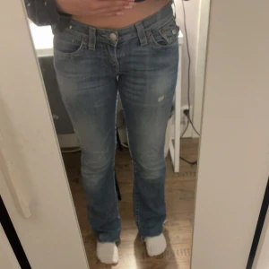True Religion ljusblå bootcut jeans - Säljer ett par ljusblå jeans från True Religion med klassiska broderade bakfickor och snygga detaljer. Jeansen har låg midja, bootcut passform och är gjorda i mjukt denim. De sitter snyggt och är väldigt lågmidjade. De är i bra skick. Storlek 27 passar mig som vanligtvis har S/36 på jeans.