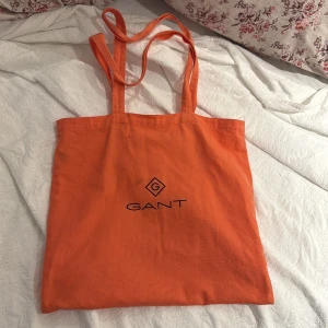 Orange shoppingväska från GANT - Orange shoppingväska från GANT i bomull med svart logga framtill. Väskan har långa handtag och ett stort huvudfack samt en innerficka med dragkedja. Perfekt för att bära med sig allt man behöver under dagen. Väl använd