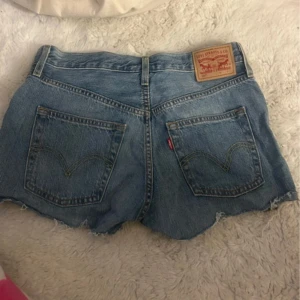 Levi's 501 jeansshorts ljusblå - Ljusblå jeansshorts från Levi's modell 501 med hög midja, klassiska fem fickor och råa, fransiga benslut. Knappar framtill och ikonisk läderpatch bak. Perfekta för en chill sommarstil.🩷