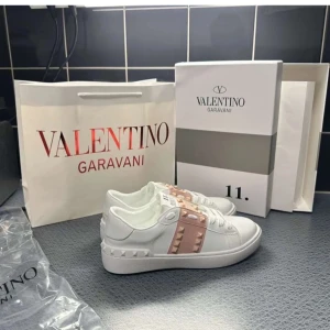Valentino Garavani Rockstud sneakers - Snygga vita Valentino Garavani Rockstud sneakers med ljusrosa band och ikoniska nitar. Skorna har rund tå, platt sula och är tillverkade i skinn. Perfekta för dig som vill sticka ut med exklusiva detaljer och modern design.