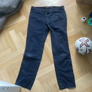 Mörkblå chinos från Stone Island - Snygga mörkblå chinos från Stone Island med raka ben och klassisk passform. Byxorna har sidofickor, bakfickor med knapp och bälteshällor. Tillverkade i bomull för en skön känsla och stilren look. Byxorna är även lagade inuti. 