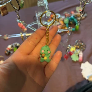 Nyckelring med grön dinosaurie - Söt nyckelring med en grön dinosauriefigur som håller en boba-te. Figuren har rosa kinder och en gul mage, och hänger i en guldfärgad kedja. Perfekt accessoar för att piffa upp dina nycklar eller väska.