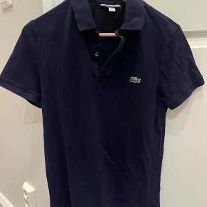 Mörkblå pikétröja från Lacoste - Klassisk mörkblå pikétröja från Lacoste med korta ärmar och den ikoniska gröna krokodilloggan på bröstet. Tröjan har krage, knappar framtill och är tillverkad i mjuk bomull. Perfekt för en clean och sportig stil.