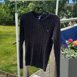 Svart kabelstickad tröja från GANT - Kabelstickad svart tröja från GANT i stretchig bomull. Klassisk rund halsringning, diskret broderad logga på bröstet och ribbade muddar. Perfekt för lager-på-lager och snygg till både jeans och kjol.