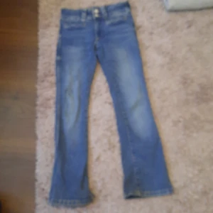 Blå bootcut jeans med dubbla knappar - Säljer ett par klassiska blå jeans med bootcut passform och snygga slitningar. Jeansen har dubbla knappar i midjan, bälteshällor och bakfickor med lock och knapp. Perfekta för dig som gillar en retro vibe och vill ha ett par jeans som sticker ut lite extra.