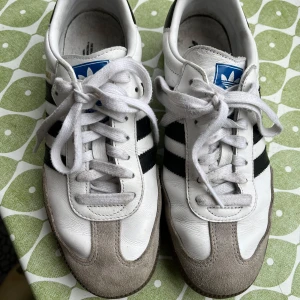 Adidas Samba sneakers vit/svart strl 40 - Klassiska Adidas Samba sneakers i vitt skinn med svarta ränder och grå mockadetaljer på tån. Skorna har vit snörning, blå logga på plösen och gummisula. Perfekt retrostil med rund tå och låg profil. Tyvärr lite för stora för mig. Skorna är väldigt lite använda.   Nypris cirka 1400 kr.