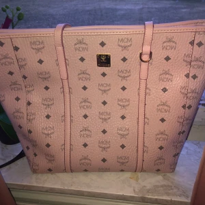 Rosa axelväska från MCM - Säljer en snygg rosa axelväska från MCM med deras ikoniska logotypmönster. Väskan har två långa handtag och en praktisk dragkedja på insidan. Perfekt för att bära dina dagliga nödvändigheter med stil.
