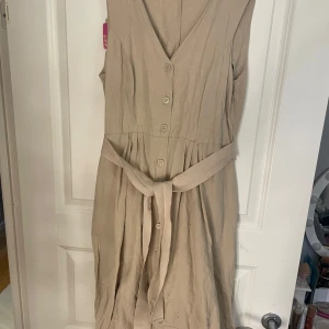 Beige ärmlös klänning med knappar - Snygg beige klänning med v-ringning och knappar framtill. Den är ärmlös och har ett knytband i midjan för en fin passform. Perfekt för varma dagar!