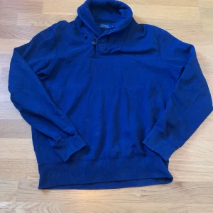 Blå tröja från Polo Ralph Lauren - Säljer en snygg blå tröja från Polo Ralph Lauren. Tröjan har långa ärmar och en hög krage med knappar. Perfekt för en stilren och avslappnad look.