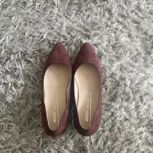 Bruna ballerinaskor från Ecco - Snygga bruna/vin röda ballerinaskor från Ecco med spetsig tå. Perfekta för en stilren look. Tillverkade i mocka för en lyxig känsla.