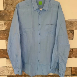 Ljusblå skjorta från Boss - Snygg ljusblå skjorta från Boss med klassisk krage och knappar framtill. Skjortan har en bröstficka och är perfekt för en stilren look. Märkeslogga på insidan av kragen.