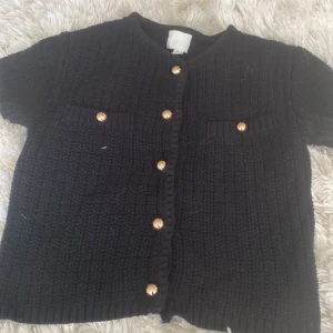 Svart stickad kofta från H&M - Säljer en svart stickad kofta från H&M med korta ärmar och guldknappar framtill. Koftan har två dekorativa fickor och en ribbad kant⭐️