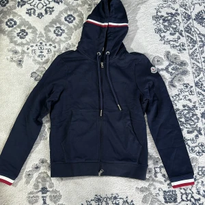 Mörkblå huvtröja från Moncler - Säljer en snygg mörkblå huvtröja från Moncler med dragkedja och ribbade muddar. Tröjan har en broderad Moncler-logga på ärmen och vita samt röda detaljer på huvan och ärmsluten. Perfekt för en stilren look!