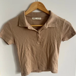 Beige ribbad polotopp från Pull&Bear - Beige ribbad polotopp från Pull&Bear. Toppen har korta ärmar och en klassisk krage med knappar framtill.