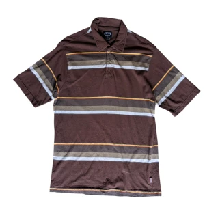 Brun randig pikétröja från Stussy - Säljer en brun pikétröja från Stussy med coola ränder i beige, vit och gul. Tröjan är tillverkad i 100% bomull och har korta ärmar. Perfekt för en avslappnad stil! Tröjan har storlek L men passar som lite större