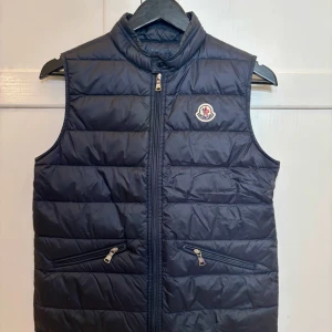 Marinblå dunväst från Moncler 14Y - Jättefin marinblå Moncler duväst barn.  Storleken är 14Y men den är lite liten i storleken. Väldigt fint skick, ser helt ny ut men den har två mindre lagningar (lagad av skräddare) på två ställen (bak till vänster nedanför kragen och framtill längst ner på vänster sida). Detta syns bara om man kollar riktigt nära. Inköpt i januari 2024 för 4 010kr och kvitto finns. 