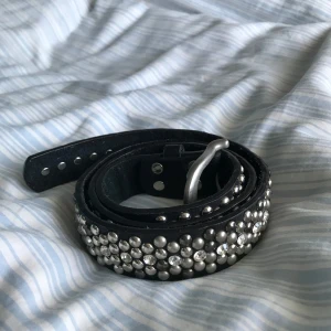 Rhinestone bälte - Snyggt svart bälte dekorerat med silverfärgade nitar och rhinestones. Tillverkat i konstläder och har ett klassiskt spänne i silver. Perfekt för att ge en edgy touch till din outfit. Storlek:85cm